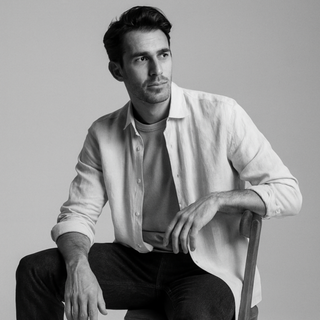 Man sitting on a chair wearing a light shirt, dark pants, and black shoes against a gray background